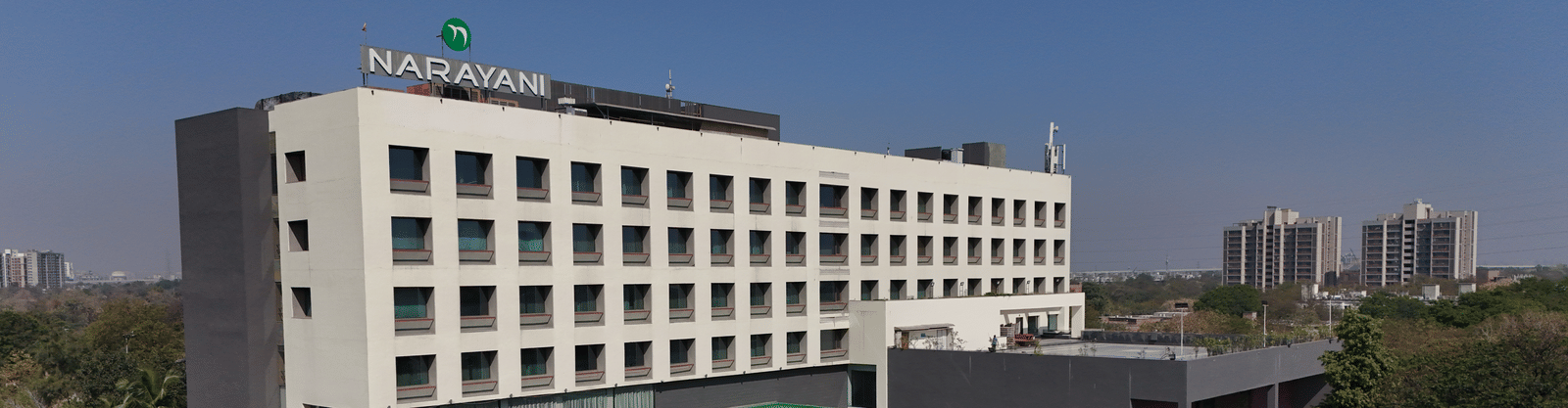 Long-shot of Narayani Heights Hotel & Resorts, Ahmedabad facade with trees surrounding it under a clear sky.