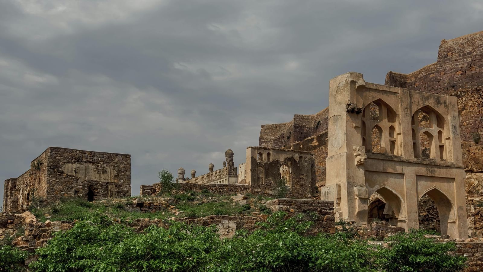 The crumbling stone ruins of Golconda Fort in Hyderabad, India, featuring arched doorways and a mosque with visible minarets, under a cloudy sky.