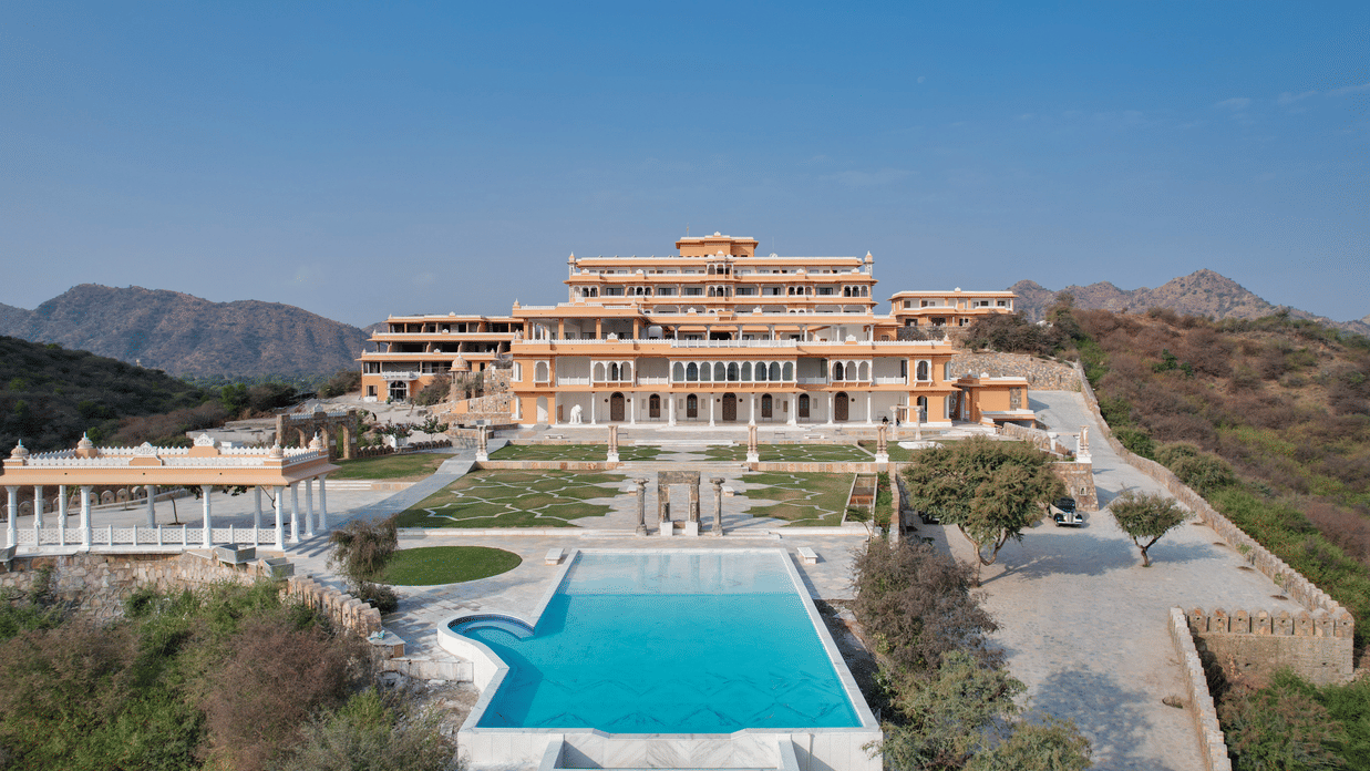 Palace-style hotel exterior with a central swimming pool, landscaped gardens, stone pathways, pavilion structures, and surrounding hills under a clear blue sky - Fateh Vilas, Udaipur