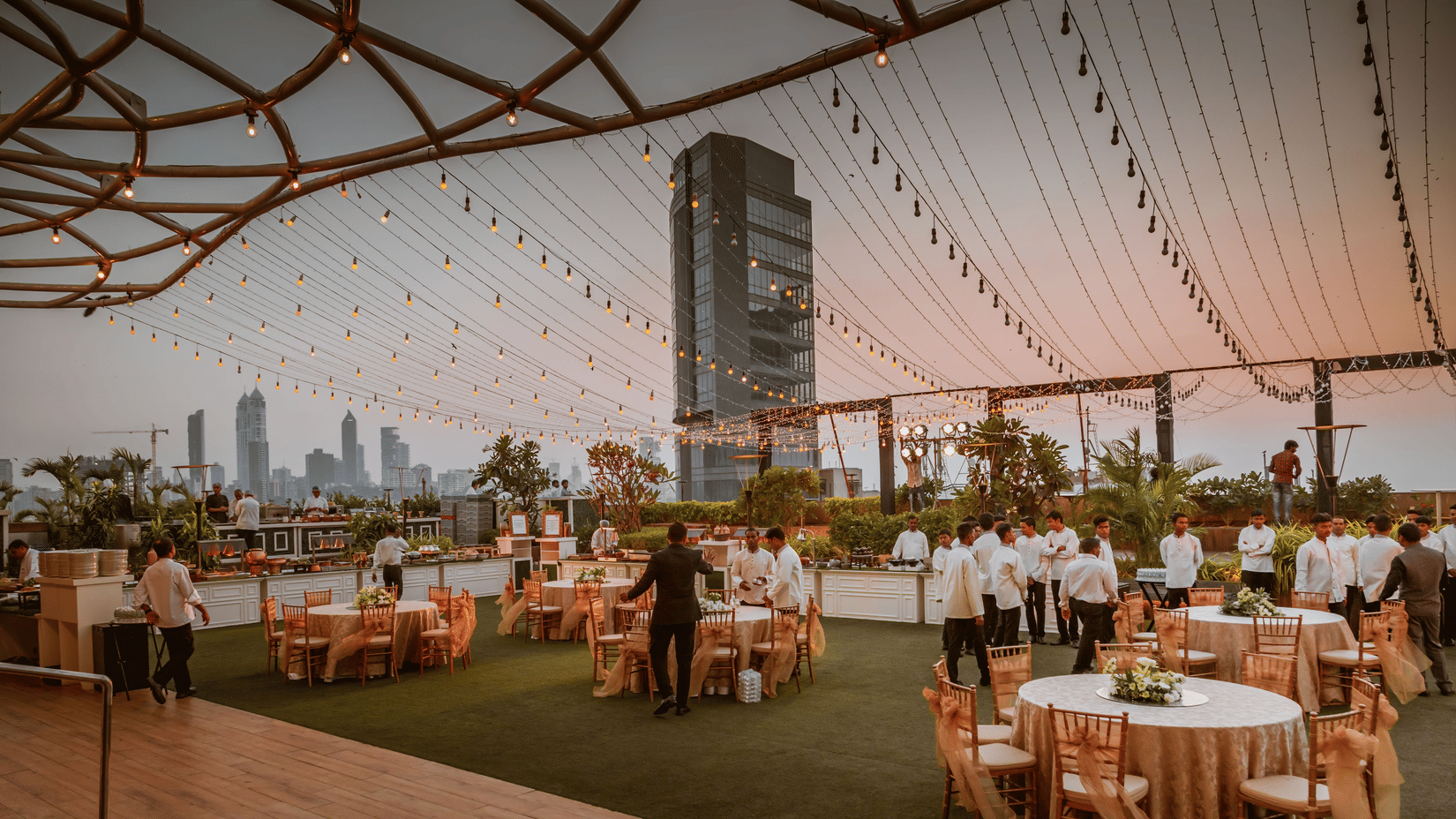 Round tables are arranged on an outdoor rooftop terrace with a mesh canopy overlooking a cityscape with skyscrapers.