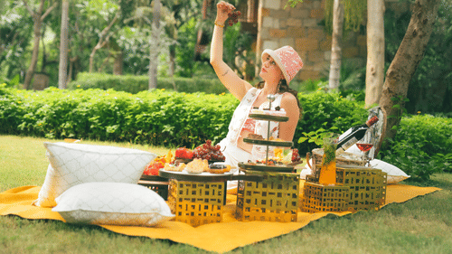 Woman enjoying an outdoor picnic on a yellow blanket in a garden setting, with cushions, a tiered tray of snacks, fruits, and drinks arranged neatly on low tables, surrounded by lush greenery - Ananta Spa and Resort, Pushkar.