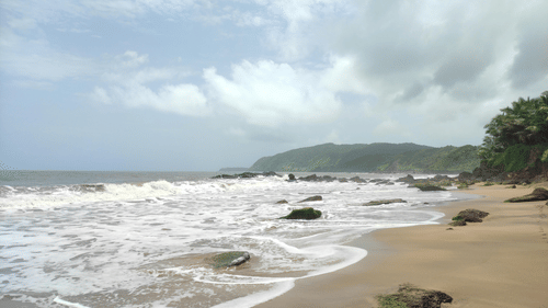 Foamy white waves crashing onto a sandy beach with rocky outcrops, set against a backdrop of green hills and grey sky.
