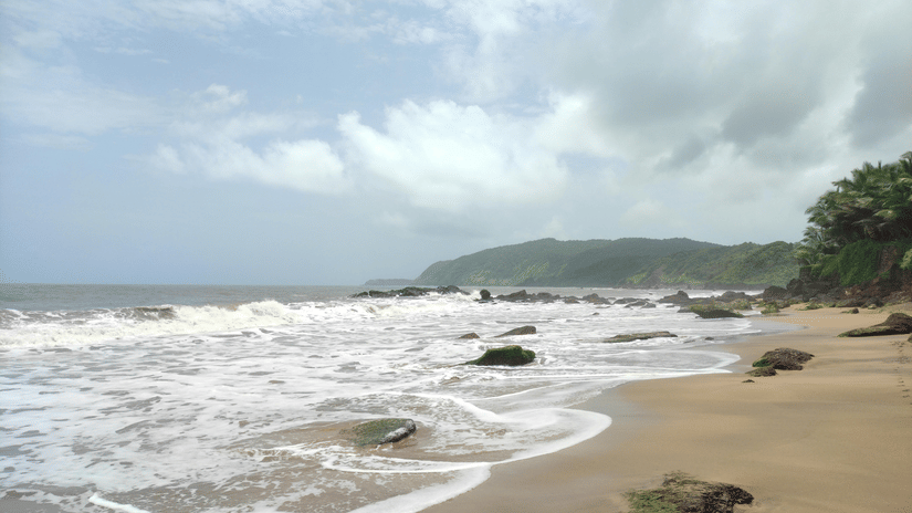 Foamy white waves crashing onto a sandy beach with rocky outcrops, set against a backdrop of green hills and grey sky.