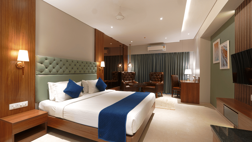 A room featuring a king size bed with a green headboard, a wooden desk, a television, and a brown leather armchair by the window at Magnus Kapitol, Sakore Nagar, Viman Nagar.