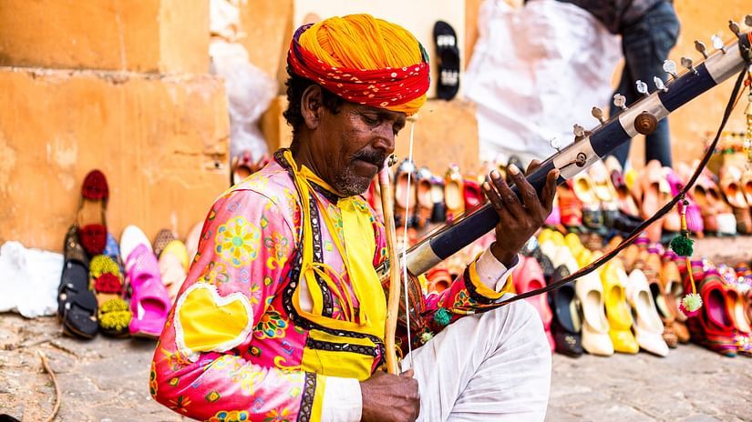 A person wearing a turban and patterned clothing plays a wind instrument while seated, with shoes displayed in the background.