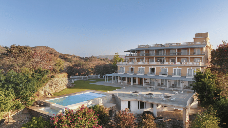 A facade of Fateh Safari Resort in Kumbhalgarh featuring a green lawn and a patio with tables and chairs. It also features a swimming pool set amidst the trees.