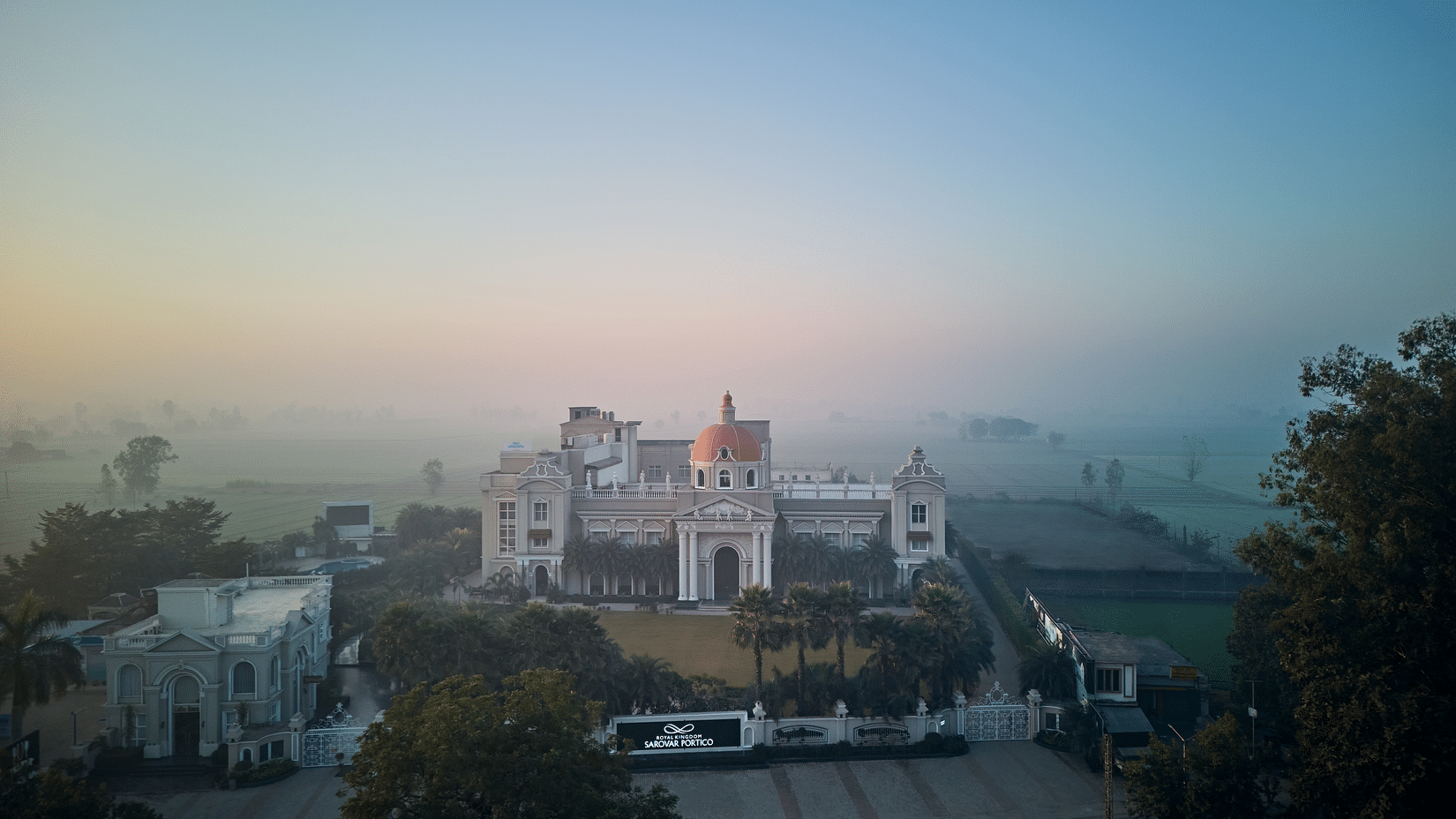 facade of Royal Kingdom Sarovar Portico, Pilibhit with fog-covered fields in the distance