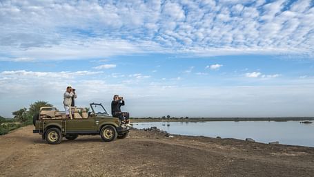 People stand in a vehicle, observing a waterside landscape under a partly cloudy sky - image provided by Fort Barli, Ajmer.
