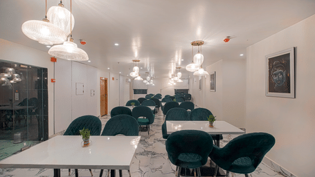 A clean, well-lit view of the modern dining room at Perfectstayz Premium Varanasi, featuring dark seating, white tables, an artistic wall frame, and marble flooring.