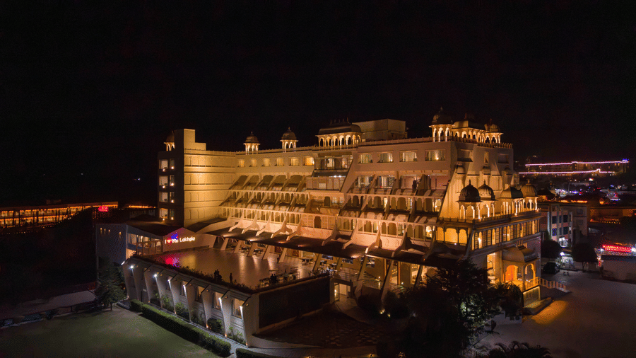 An evening drone view of Via Lakhela Lakeview Resort, highlighting the grand illuminated facade, waterfront deck, and vibrant lighting across the property.