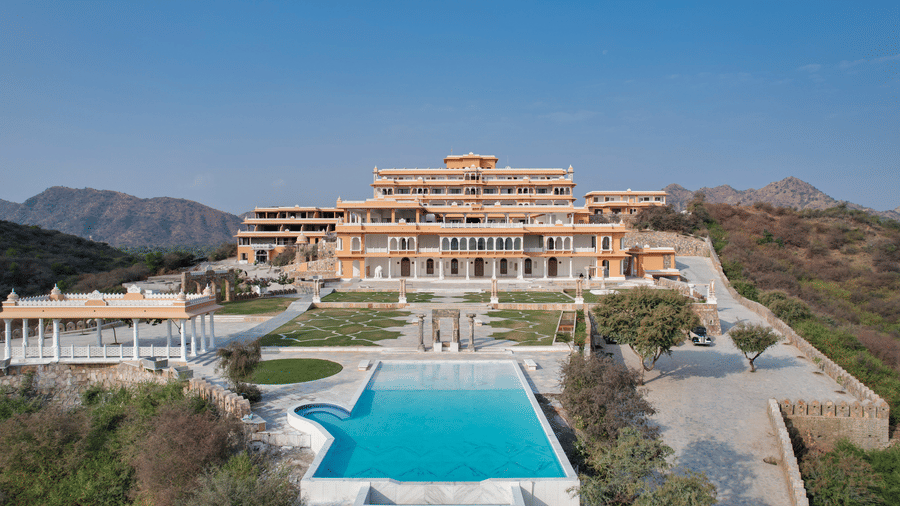 Palace-style hotel exterior with a central swimming pool, landscaped gardens, stone pathways, pavilion structures, and surrounding hills under a clear blue sky - Fateh Vilas, Udaipur