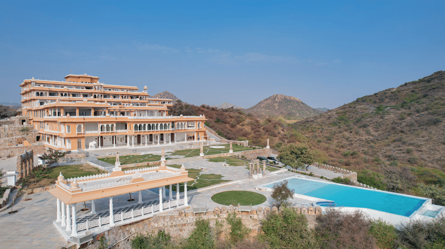 The facade of Fateh Vilas with a swimming pool in front of the property surrounded by mountains