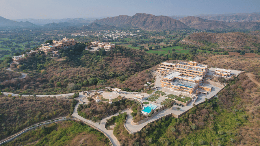 Aerial view of Fateh Vilas, the best resort in Udaipur, surrounded by green mountains with a swimming pool, a road, walking pathways, and lawns