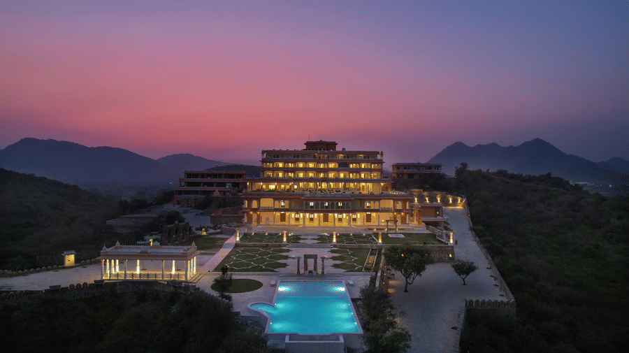 A far out view of Fateh Vilas, a heritage resort in Udaipur, during twilight hour, with a swimming pool, a large lawn area, a canopy, walking pathways, and mountain in the background seen.