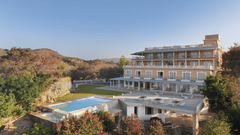 A facade of Fateh Safari Resort in Kumbhalgarh featuring a green lawn and a patio with tables and chairs. It also features a swimming pool set amidst the trees.
