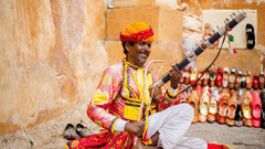 A rajasthani folk person sitting on the ground next to a stone wall holding his music instrument.