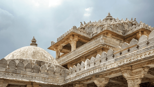 Intricately carved marble temple exterior showcasing ornate domes, decorative pillars, detailed cornices, and traditional Jain architecture beneath a cloudy sky.
