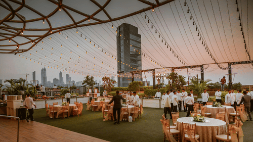An outdoor event space under a large canopy, featuring dining tables and chairs, with a city skyline visible in the background.