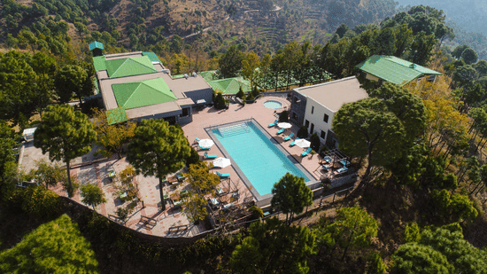 a large bird-eye shot of Suryavilas Luxury Resort and Spa featuring its swimming pool and premises surrounded by lush greenery on a hilltop