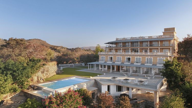 A facade of Fateh Safari Resort in Kumbhalgarh featuring a green lawn and a patio with tables and chairs. It also features a swimming pool set amidst the trees.