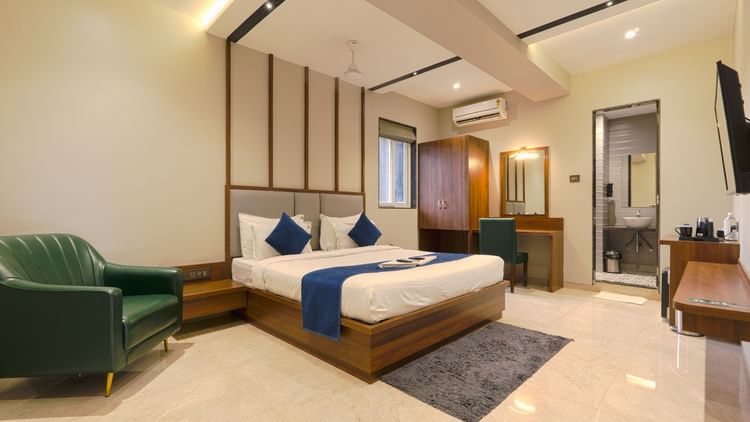 A spacious, modern hotel room featuring a wooden double bed with white linens and blue accents, a green armchair, a work desk, wardrobe, wall-mounted TV, and an attached bathroom - Magnus Fortune House, Magarpatta.