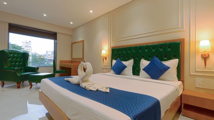 A well-lit hotel room featuring a large bed with a green tufted headboard, blue pillows and runner, towel swans on the bed, green armchairs by a window, and warm wall-mounted lamps - Magnus Nexstar Suites, Kalyani Nagar