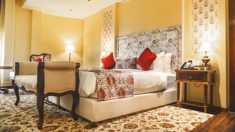 The energised look of the Super deluxe cottage at The Garden Ananta Elite with enchanting white table lamps on both sides, red pillows to up the comfort, and a cushioned seat for two, placed adjacent to the bed.