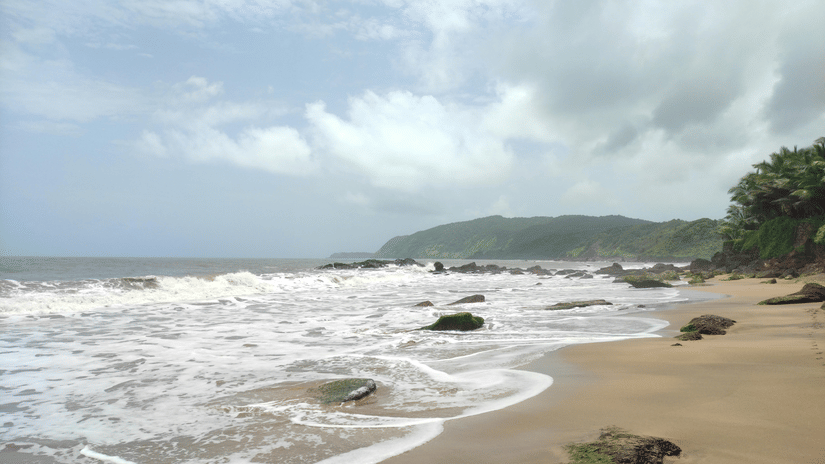 Foamy white waves crashing onto a sandy beach with rocky outcrops, set against a backdrop of green hills and grey sky.