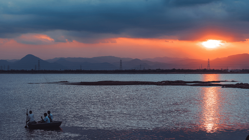 A small fishing boat drifts across calm waters as the setting sun casts orange reflections beneath dramatic evening clouds and hills