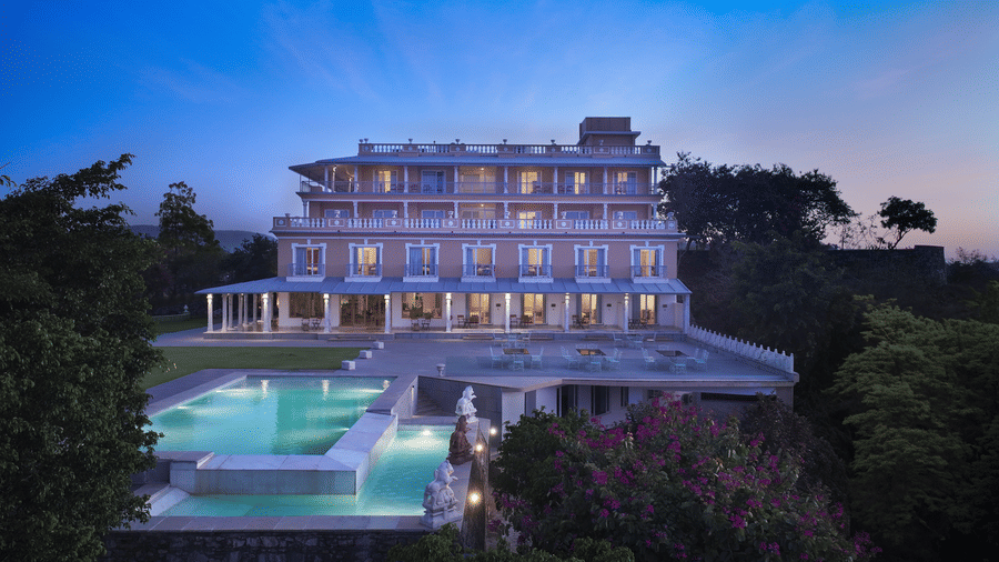 A facade of Fateh Safari Resort, Kumbhalgarh featuring four storey building, a swimming pool, a patio for sitting area with tables and chairs, and a green lawn.