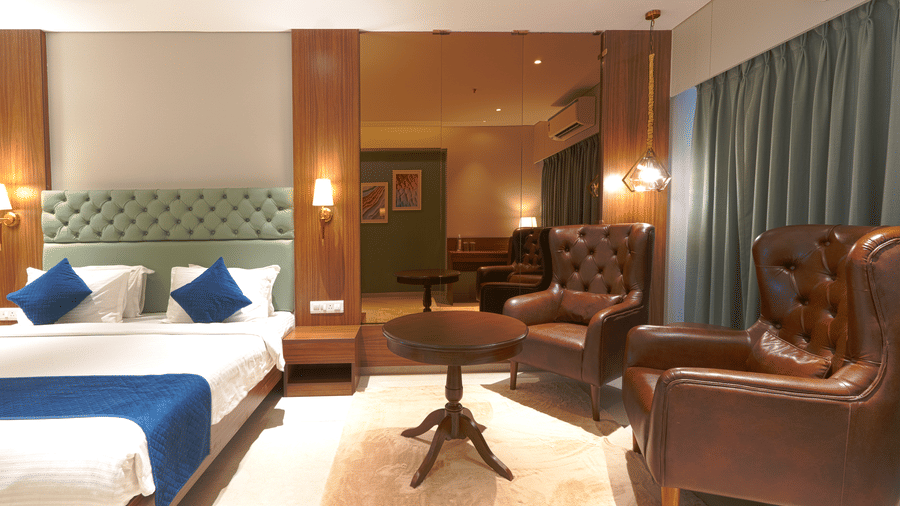 A room with a bed featuring green headboard, white bedding with a blue runner, a small round table, and a brown leather armchair at Magnus Kapitol, Sakore Nagar, Viman Nagar.