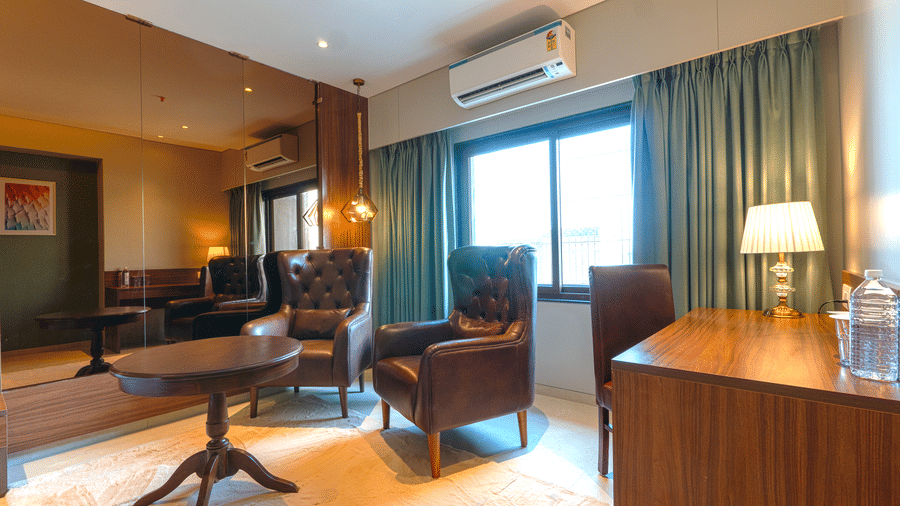 A seating area featuring a round wooden table, 2 leather armchairs, a desk with a lamp, and teal curtains by a window at Magnus Kapitol, Sakore Nagar, Viman Nagar.
