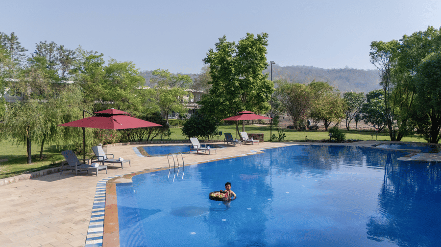 Swimming pool with a person in an inflatable ring, surrounded by a paved deck, green trees  at The Golden Tusk, Jim Corbett