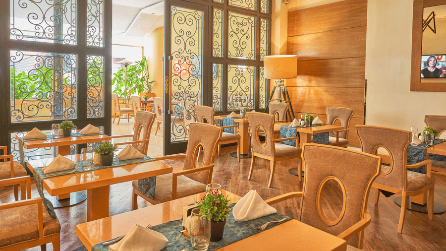 Restaurant dining area with wooden tables and chairs, folded napkins, placemats, potted plants as centerpieces, large decorative glass windows, and warm inviting ambiance.