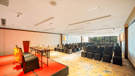 Godavari banquet hall in Vijayawada with a stage, podium, theatre-style seating, and floor-to-ceiling windows, set up for a corporate conference or seminar | Grand Vijayawada by GRT Hotels