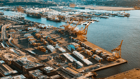 An aerial view of a sprawling seaport terminal with neat rows of cargo and containers, bright yellow cranes, and ships docked across the harbour.