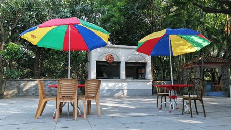Two outdoor dining tables with chairs under bright, multicoloured umbrellas next to a small white kiosk.