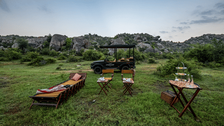 An outdoor luxury safari camp setup with lounge chairs, small tables, and a vehicle in a grassy area surrounded by lush green hills.
