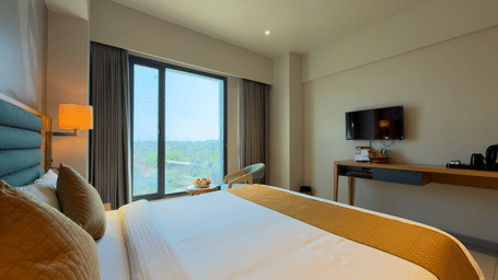 Hotel deluxe room with bed side tables, wall art, television, chair, desk, curtains, and window showing outdoor view