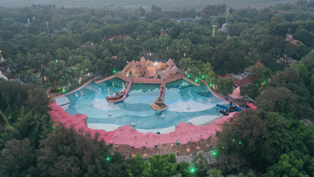 An aerial view of a wave pool inside Water Kingdom surrounded by many trees as seen during late evening time with the lights switched on. 