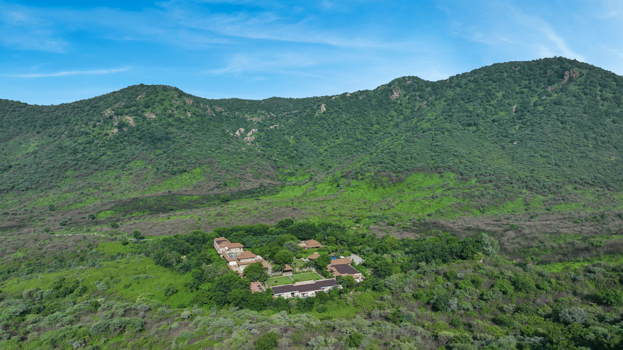 A distant view of the resort buildings nestled at the base of a green mountain | Anandam Jungle Retreat Resort, Udaipur