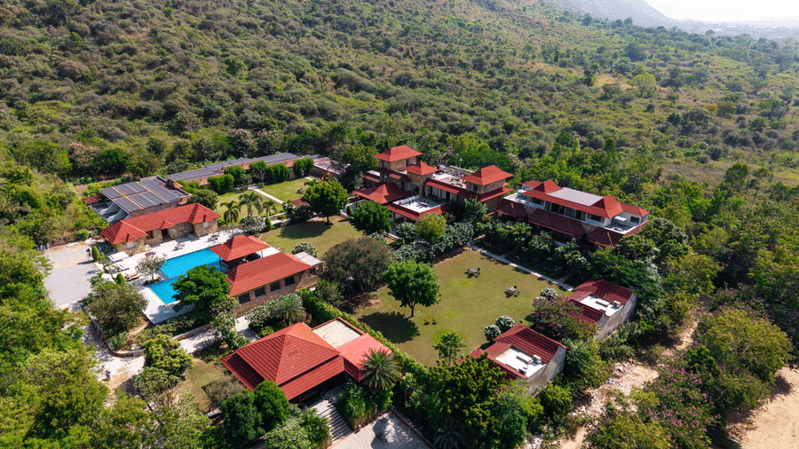 An aerial view of a resort complex with red-roofed buildings and a swimming pool surrounded by forest | Anandam Jungle Retreat Resort, Udaipur