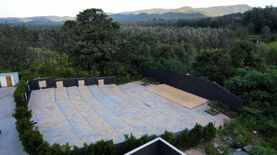 An aerial view of a large, level outdoor area, partially paved with pale blue tiles or mats, surrounded by dense green forestry and hills under a clear sky at Bindiga Peak Resorts.
