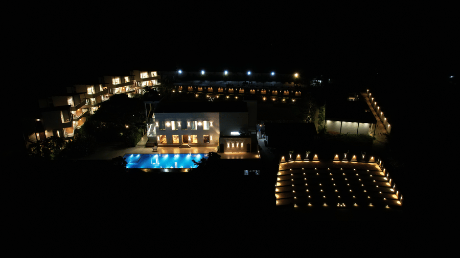 A brightly lit swimming pool, main building, and surrounding pathways against a dark background at Bindiga Peak Resort.