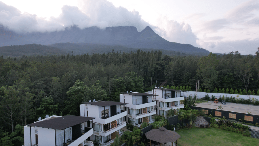 White resort buildings next to a dense forest with a large mountain covered in low clouds behind at Bindiga Peak Resort.
