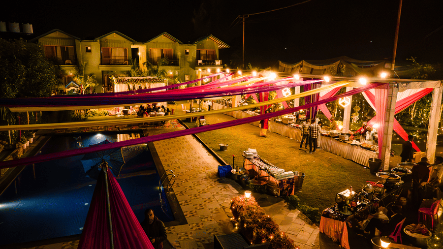 Night-time view of an outdoor event area at Corbett Nirvana Resort, showing long pink and yellow drapes strung across the open space and various light sources.
