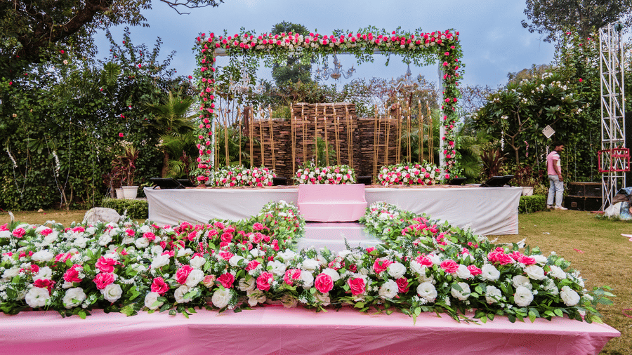 Day-time view of a wedding stage setup on a lawn at Corbett Nirvana Resort, featuring a backdrop made of wooden frames and lush pink and white floral arrangements.