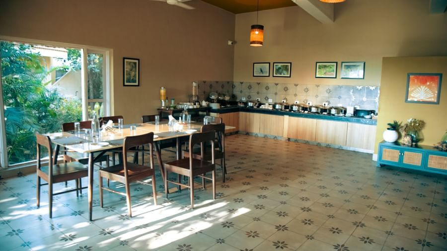 Indoor dining area with neatly arranged tables and chairs, wall decor, and natural light entering through large windows at Fireflies Resorts, Kabini.