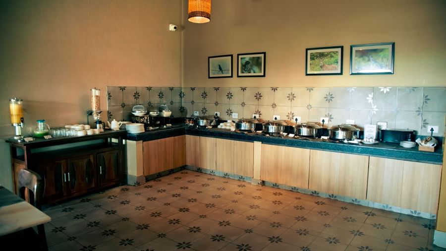 Breakfast buffet with chafing dishes and dispensers lined along a corner counter in a tiled room at Fireflies Resorts, Kabini.