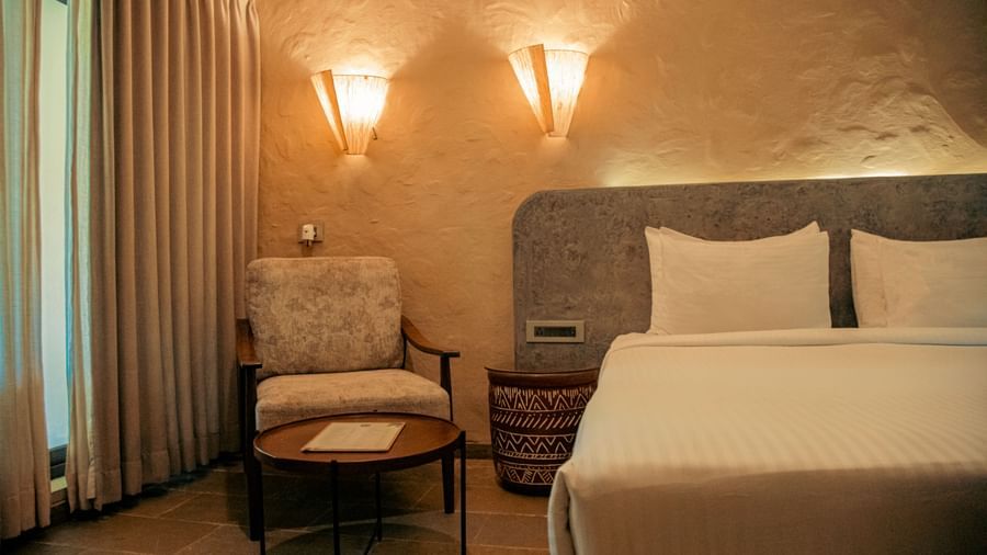 A partially visible bed with padded headboard, flanked by reading lights and a chair beside the window at Fireflies Resorts, Kabini.